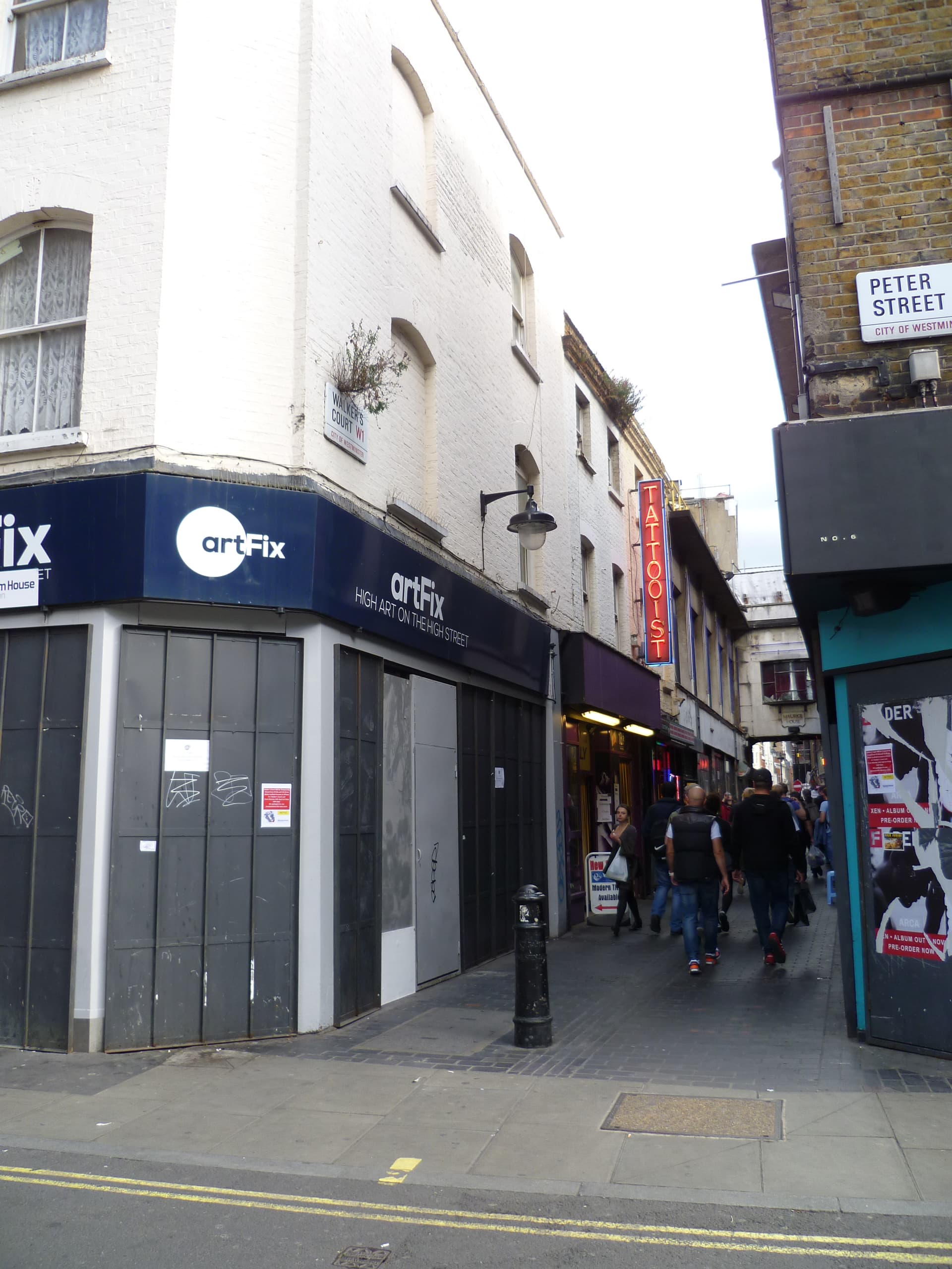 Narrow Soho alley off Peter Street with a neon tattooist sign, shuttered corner shop and pedestrians walking along