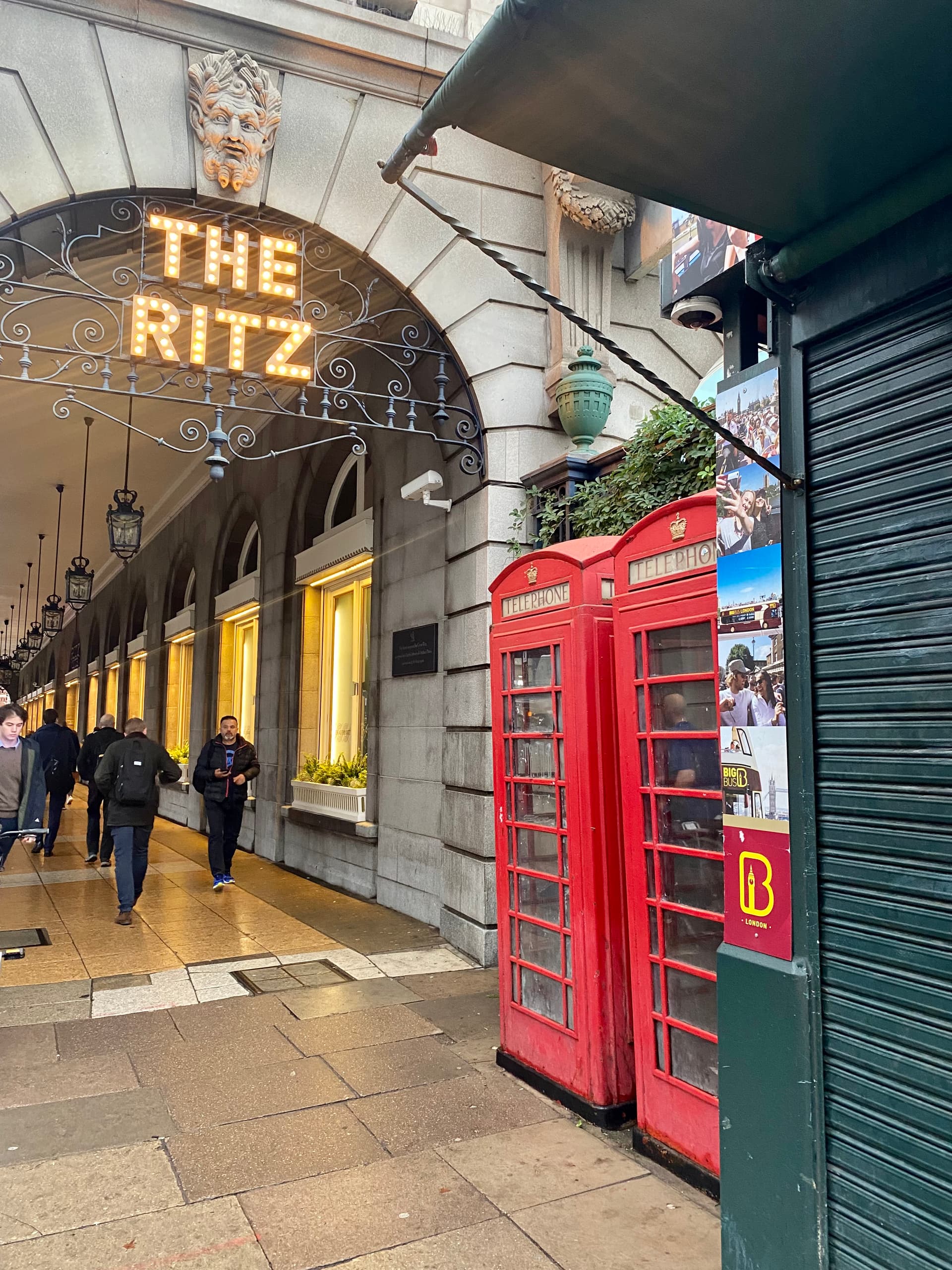 Illuminated Ritz sign above the Piccadilly arcade, with pedestrians walking past and two red telephone boxes beside it