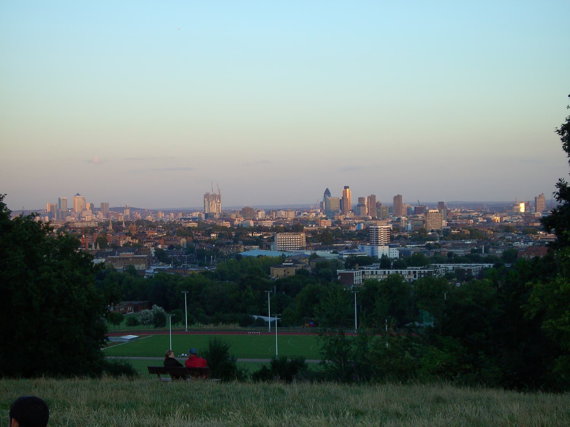 Evening view across London's skyline from a grassy hilltop, with the Gherkin and Canary Wharf beyond trees and playing fields