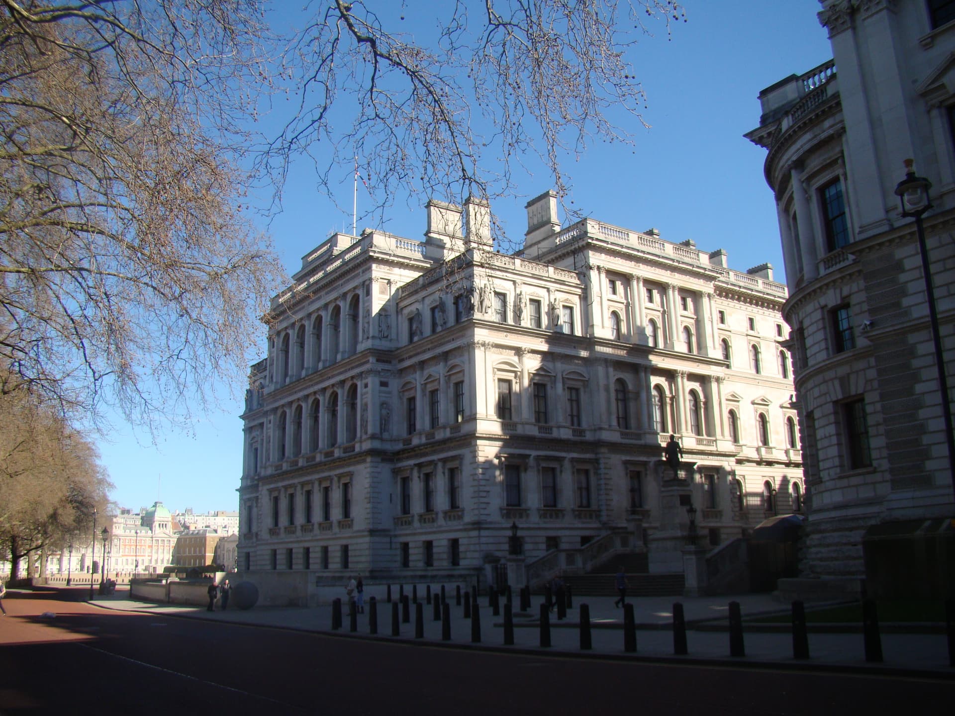 Sunlit Portland stone government building in Westminster seen through bare plane tree branches, with bollards along the pavement