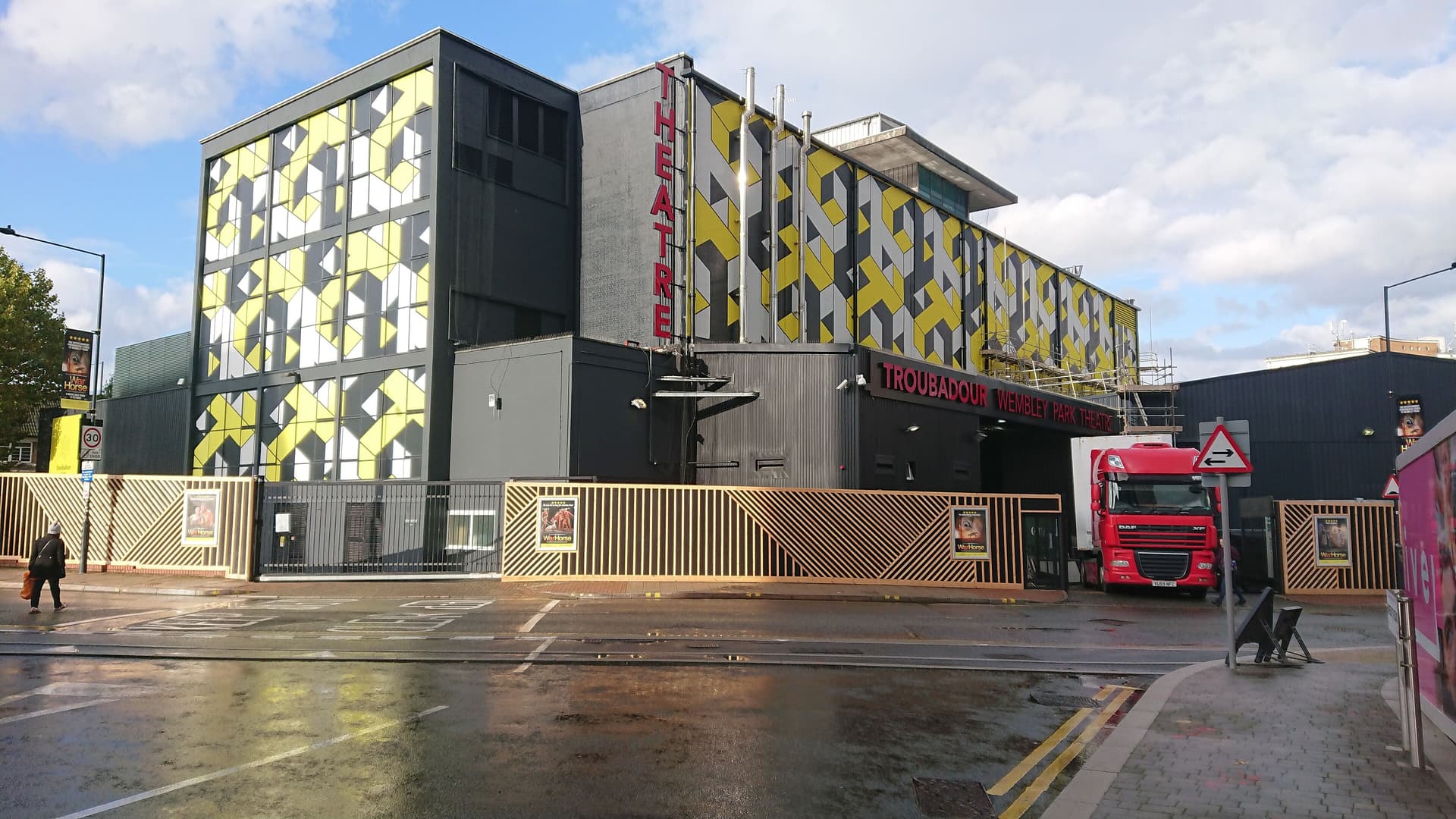 Troubadour Wembley Park Theatre's yellow, grey and black geometric facade beside a wet street, with a red lorry outside