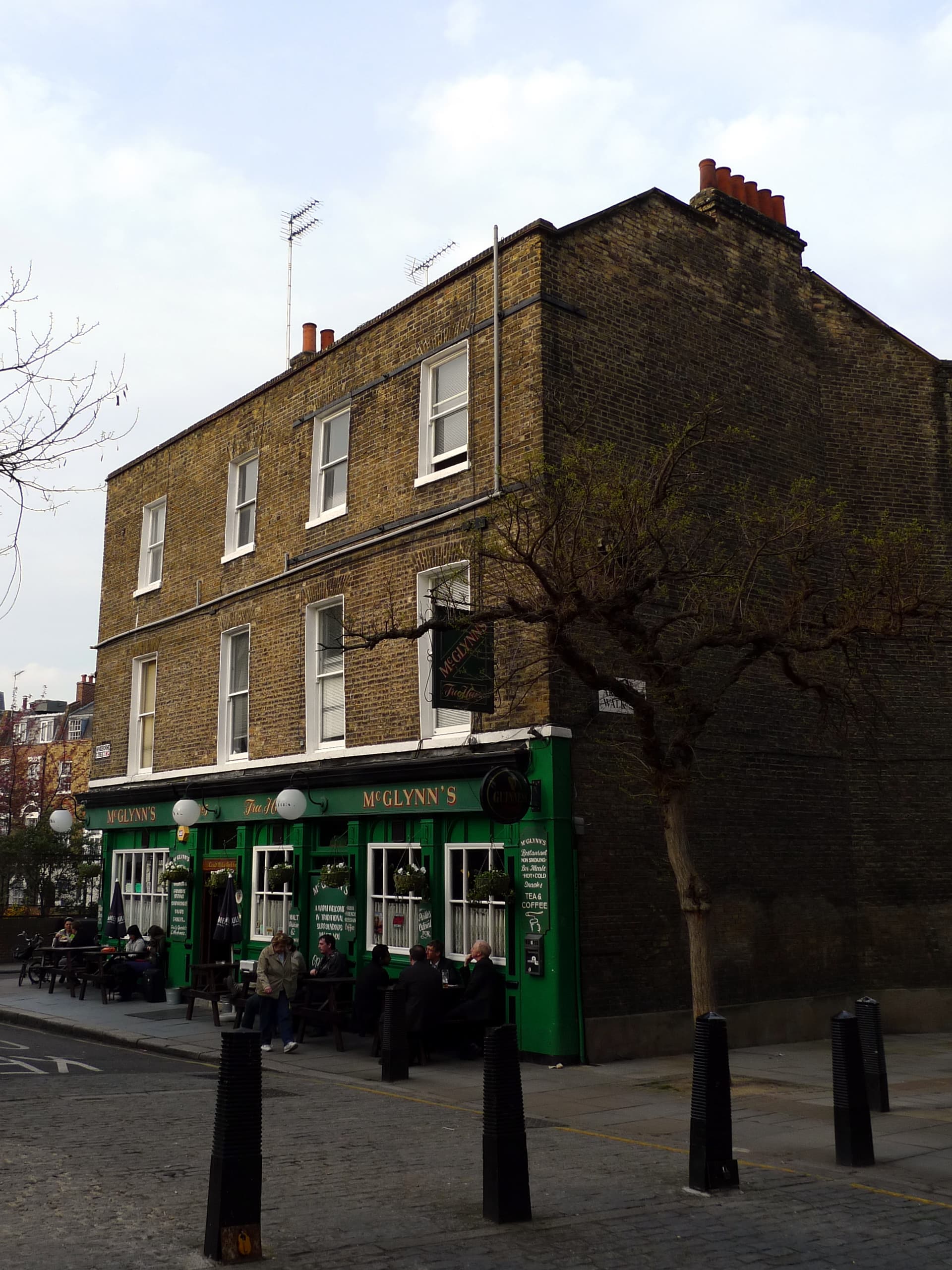 Green-fronted McGlynn's pub on a corner of yellow London brick, with drinkers seated outside and black bollards in front