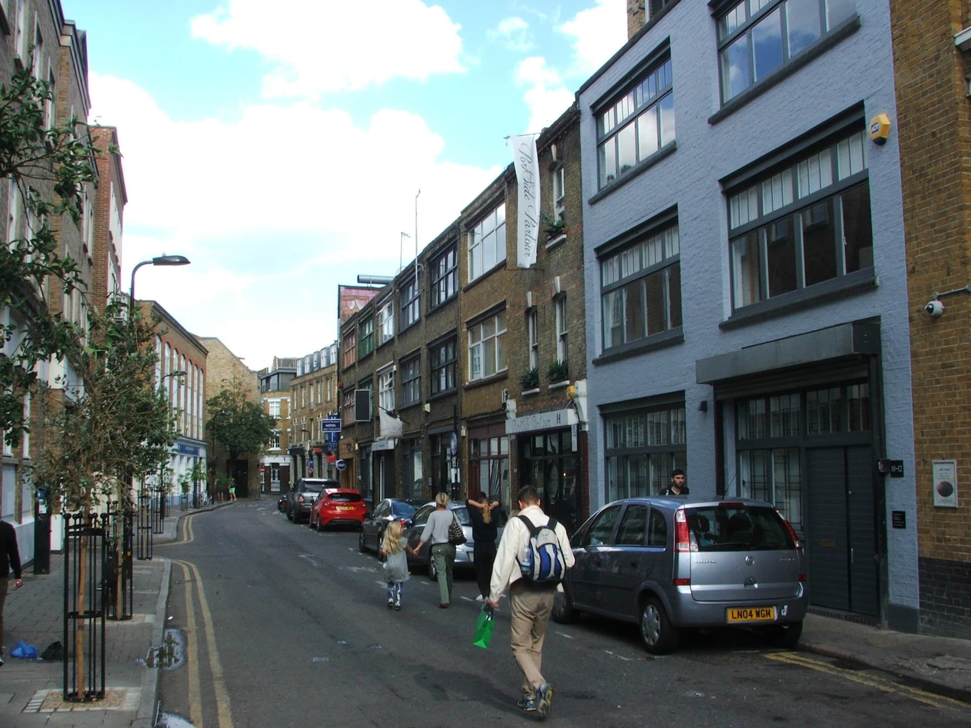 Pedestrians walk along a Shoreditch street lined with brick warehouses, parked cars and a painted lilac building under blue sky