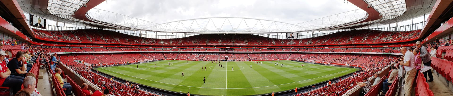 Panoramic view inside Emirates Stadium, with red seating tiers, a striped pitch and players warming up