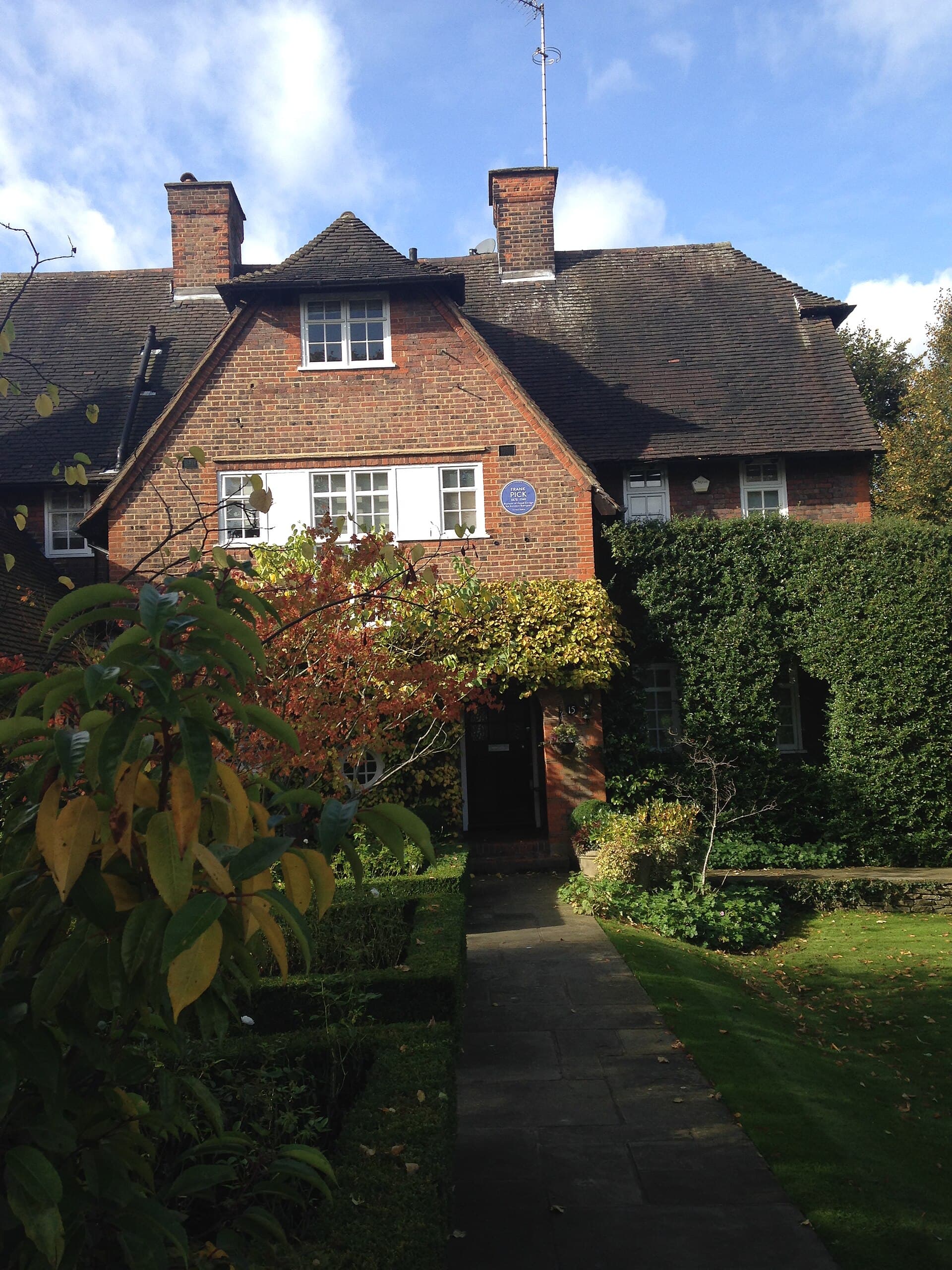Red-brick Arts and Crafts house with blue plaque for Frank Pick, set behind autumn garden planting and a flagstone path