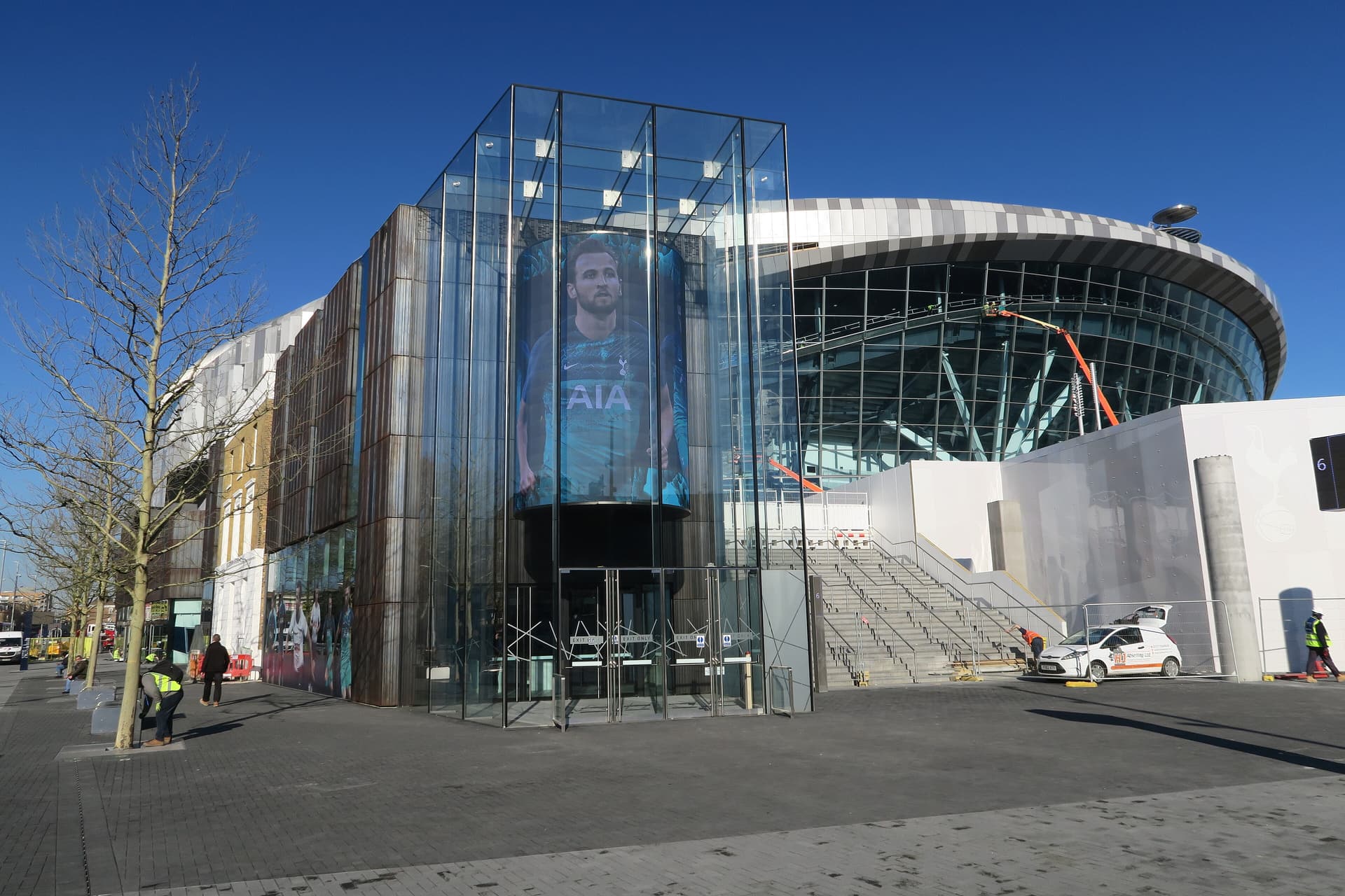 Glass entrance pavilion and curved stand of Tottenham Hotspur Stadium under a clear blue sky, with workers on the plaza
