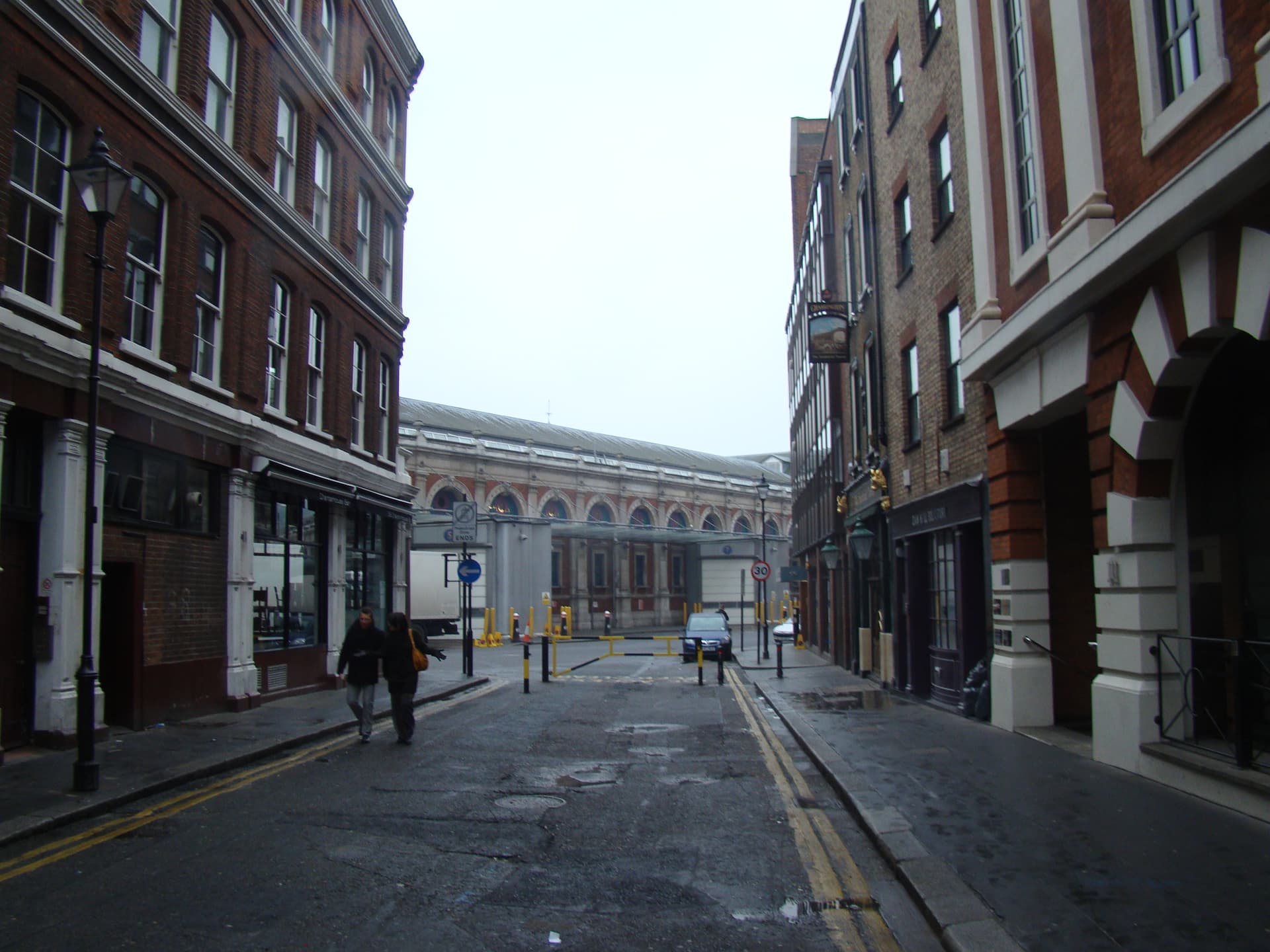 Wet brick-lined London street leading to a barrier and the arched, red-brick Smithfield Market building beyond