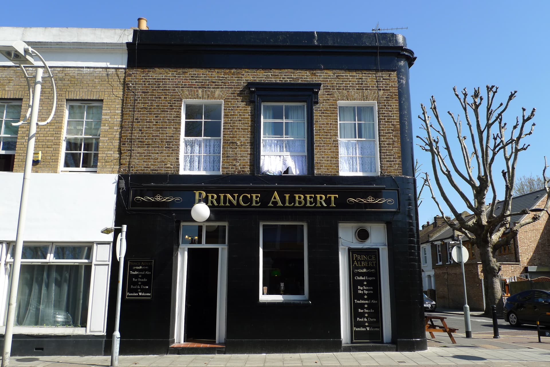 Black-tiled corner pub with gold Prince Albert signage and yellow brick upper storey on a sunlit London street