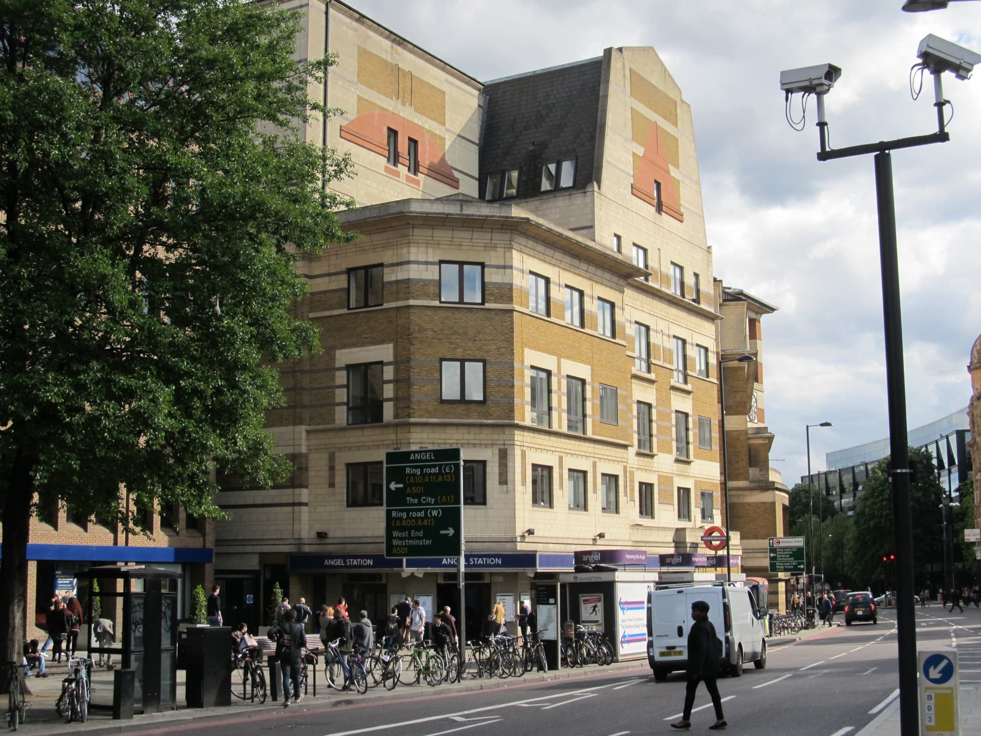 Angel Underground station entrance beneath a buff-brick office building, with bicycles, pedestrians and road signs on Islington's busy High Street