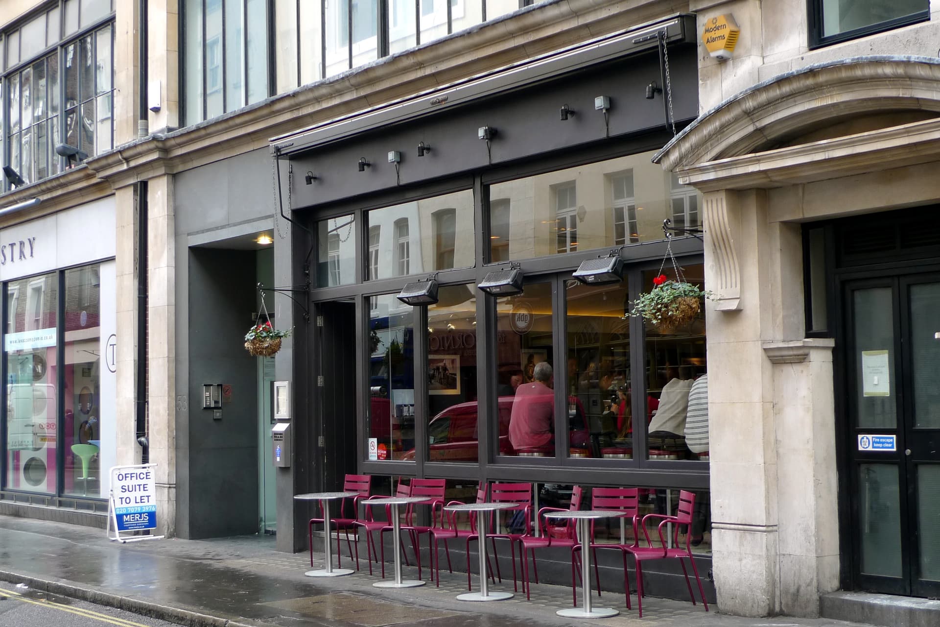 Dark-framed restaurant frontage on a wet London street with red metal chairs and tables outside and diners seen through the windows