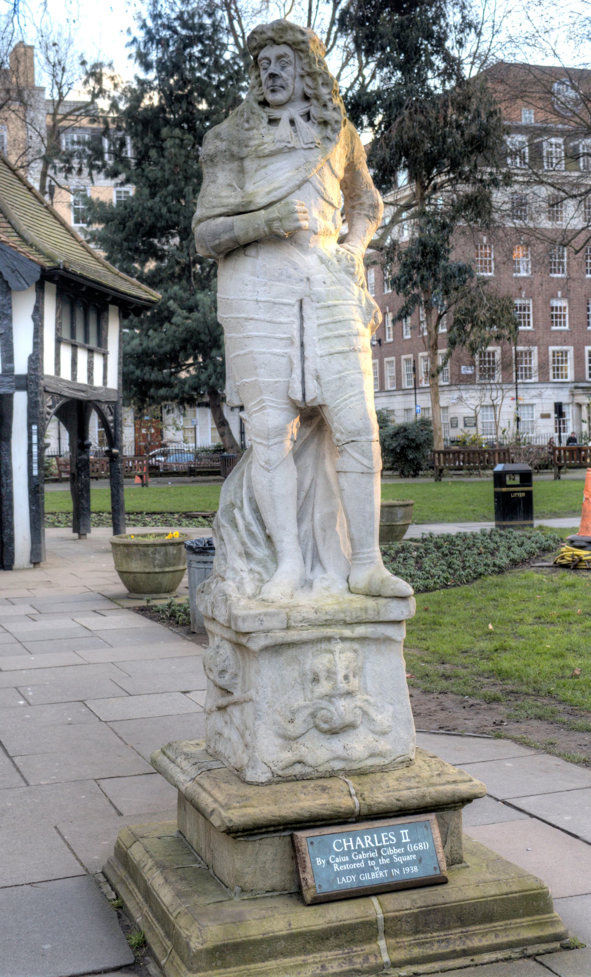 Weathered stone statue of Charles II on a plinth in Soho Square garden, with Tudor-style hut and brick buildings behind