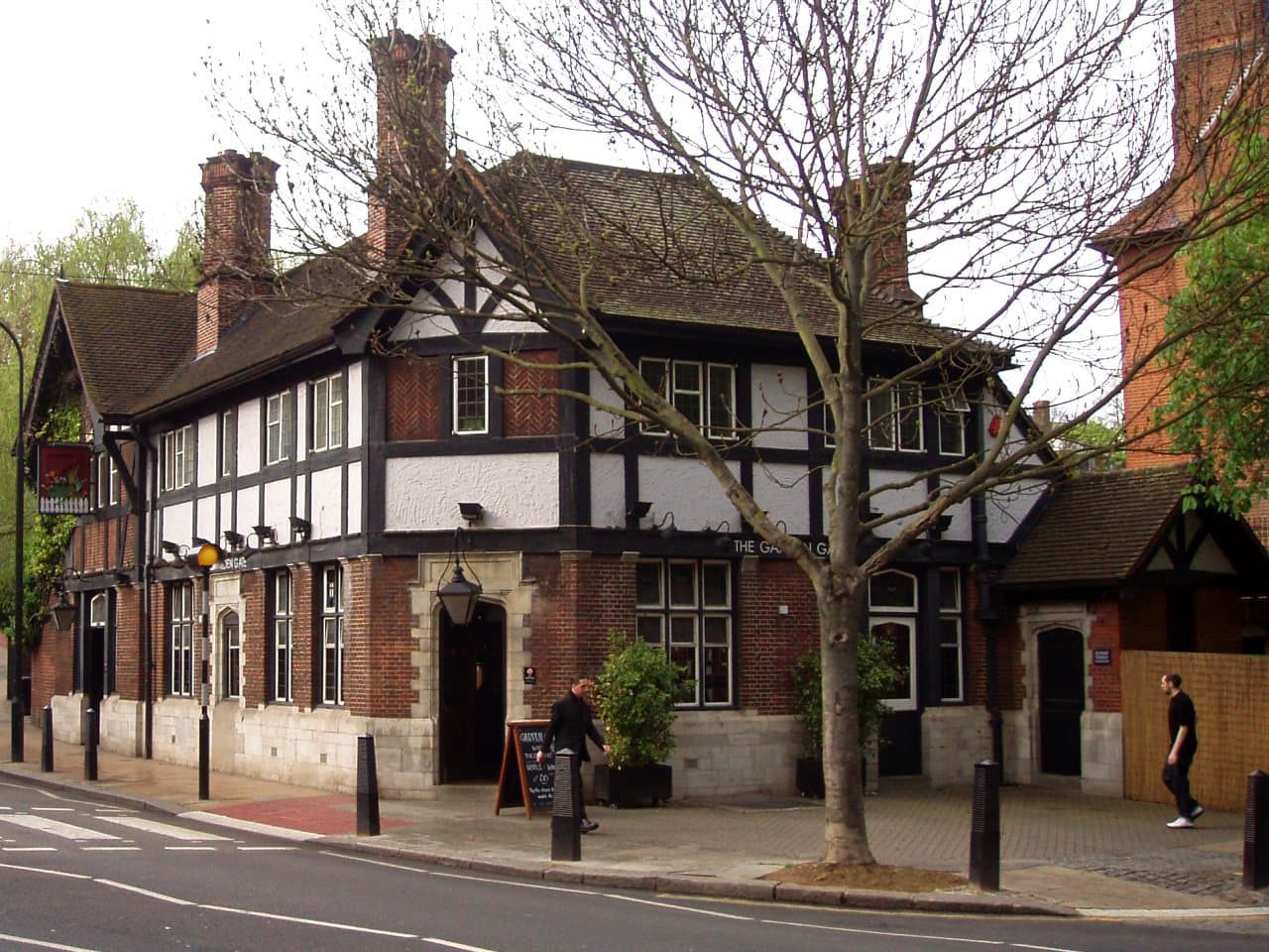 Black and white timbered corner pub with red brick ground floor, tall chimneys and a bare tree outside