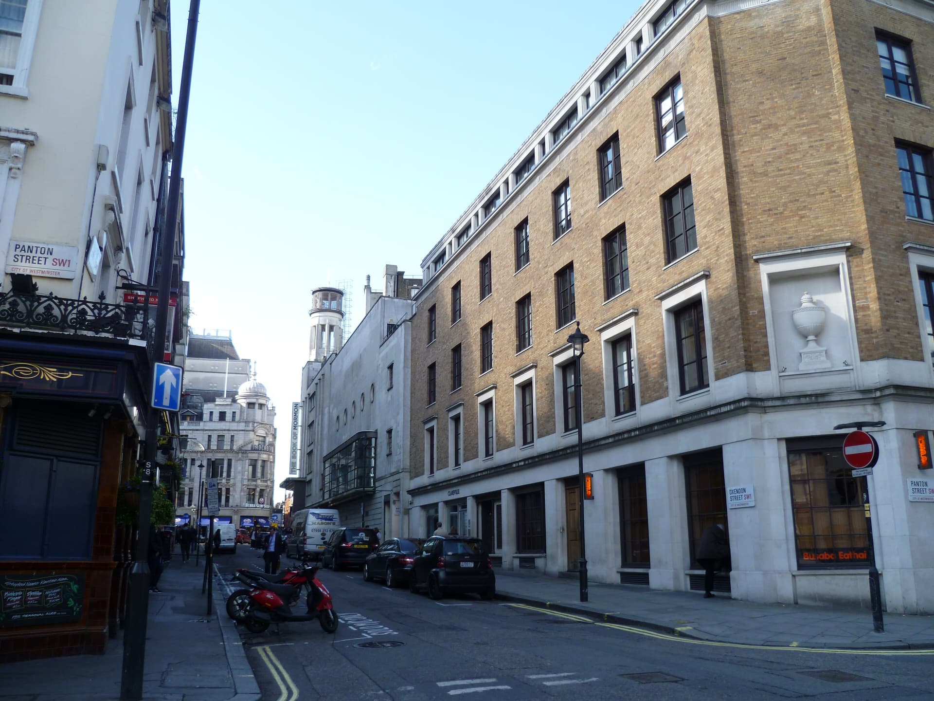 Brick and stone buildings along Oxendon Street in Soho, with parked scooters, cars and a theatre tower beyond